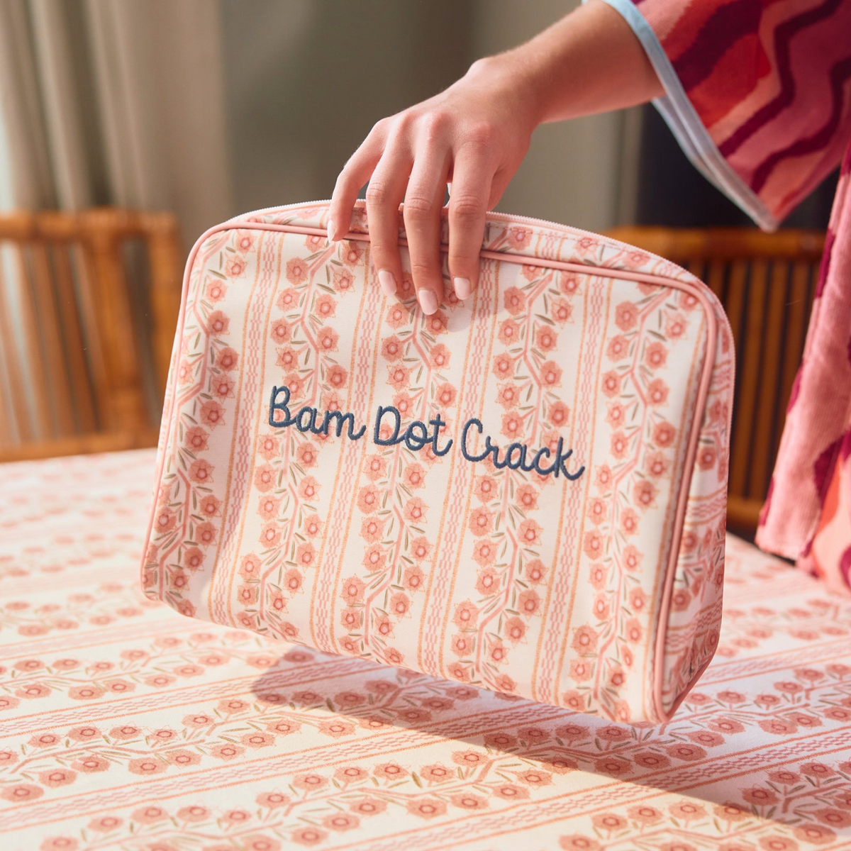 A hand holding a rectangular toiletry bag featuring a pink and coral floral stripe pattern, set against a matching patterned fabric background.