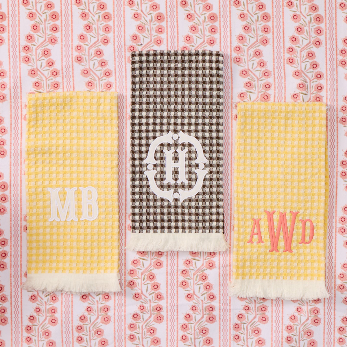 Three personalized kitchen towels laid flat on a pink and white floral patterned fabric background.